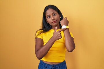 Young indian woman standing over yellow background in hurry pointing to watch time, impatience,...