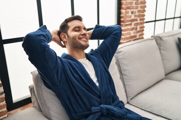 Young hispanic man wearing bathrobe relaxed with hands on head at home