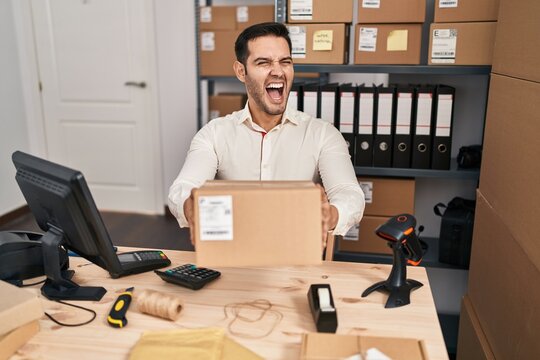 Young hispanic man with beard working at small business ecommerce holding box angry and mad screaming frustrated and furious, shouting with anger. rage and aggressive concept.