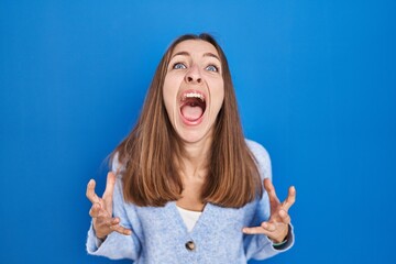 Young woman standing over blue background crazy and mad shouting and yelling with aggressive...