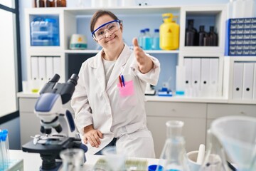 Fototapeta premium Hispanic girl with down syndrome working at scientist laboratory smiling friendly offering handshake as greeting and welcoming. successful business.