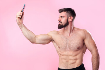 Strong and muscled bodybuilder taking a selfie with his smartphone of his shirtless torso, showing off his gains to friends on social media