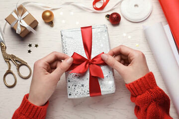 Christmas present. Woman tying ribbon bow on gift box at white wooden table, top view