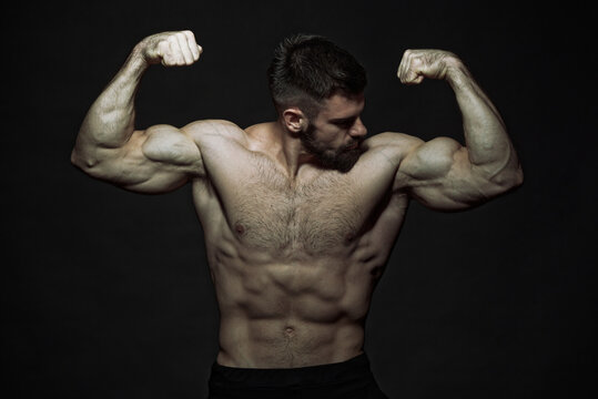 Muscly shirtless man, a bodybuilder, flexing his biceps, showing off his big arms and muscular body, dark image, black background