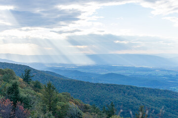 Rays of light can be seen shining through the clouds over the blue Ridge Mountains inside Shenandoah National Park. The foreground of the photo features the brush and forests of a mountain.