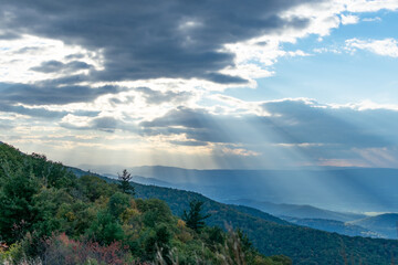 Fototapeta na wymiar Rays of light can be seen shining through the clouds over the blue Ridge Mountains inside Shenandoah National Park. The foreground of the photo features the brush and forests of a mountain.