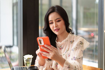 Young woman using smartphone at table with laptop in coffee shop.