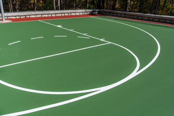Interesting green and red outdoor basketball court at school playground.  Court includes retaining walls and black vinyl coated chain link fence.	