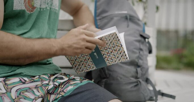 Man sitting on a bench with his backpack in a park and reading a booklet about hiking.