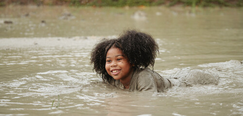Happy african-american children girl playing in wet mud puddle during raining in rainy season.