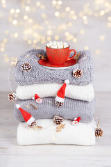 A stack of folded knitted sweaters, a red cup of cocoa with marshmallows, a garland with Christmas decorations on a background of bokeh lights. Cozy homely atmosphere of the Christmas holiday. Vertica