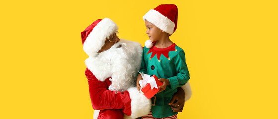 Little African-American boy in elf costume with gift and Santa Claus on yellow background