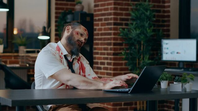 Scary looking zombie trying to work on modern laptop in office workspace. Spooky doomsday brain dead monster with deep and bloody wounds and scars trying to use portable computer in agency space.