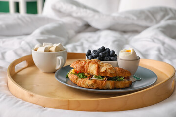 Wooden tray with tasty croissant, boiled egg, blueberry and cup of cacao on bed, closeup