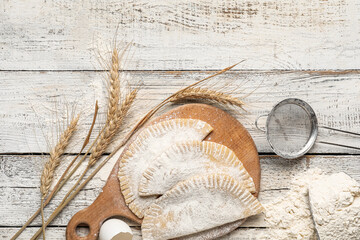Board with raw chebureks, spikelets, flour and sieve on white background