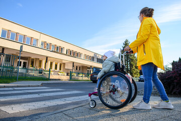 Mother pushing wheelchair with her daughter, young girl living with cerebral palsy, on their way to...