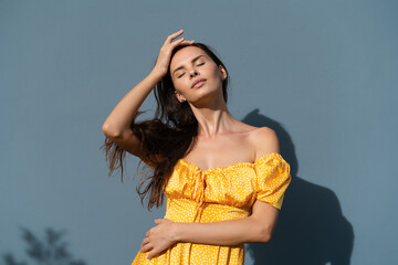 Pretty woman long dark hair in bright yellow summer dress posing on blue wall background day natural light outdoor