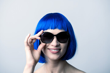 Portrait of beautiful and sexy woman with blue wig and sunglasses looking to camera with smile. Sun tan, pink lipstick and white teeth. Model with naked shoulders. Toned image with blue color