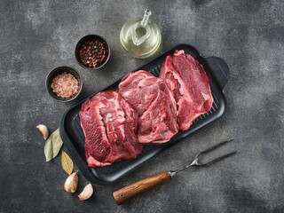 Raw pork cheeks meat on a dark gray surface with spices. Top view