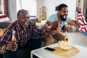 Senior father and adult son cheering for american football game at home