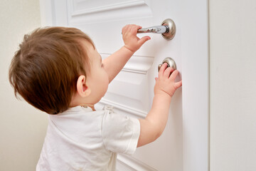 Toddler baby opens the lock, holding the door handle, child hand close-up. White wooden door, metal...