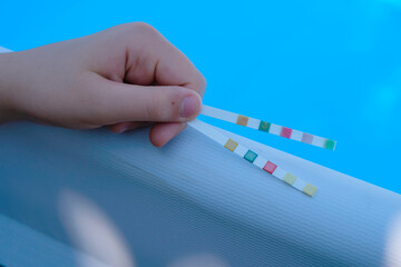 Pool test strips in the child's hand. Blue pool background. Water balance. - obrazy, fototapety, plakaty