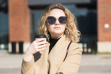 A young Caucasian girl in a coat drinks coffee and thinks while walking in the city in autumn.