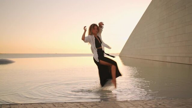 Female dancer dancing in water at sunset light. Sensual dance. Barefoot woman dressed black dancing seductive in slow motion, freedom concept. Freestyle contemporary dance