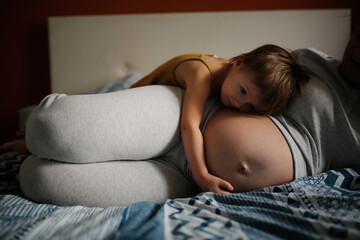 Middle-aged pregnant mother and toddler baby together on bed in bedroom in real interior.
