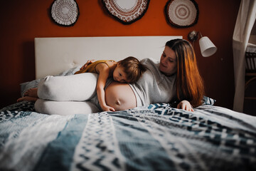 Middle-aged pregnant mother and toddler baby together on bed in bedroom in real interior.