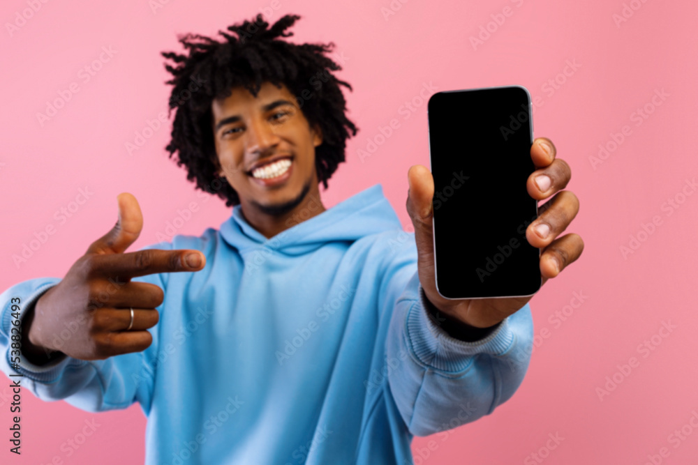 Poster happy black teen guy pointing at smartphone with mockup for website or app design on blank screen ov