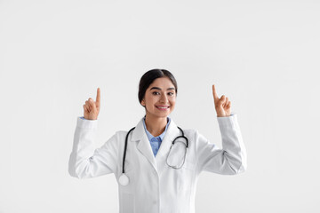 Happy millennial indian lady doctor in coat with phonendoscope raises hands and shows fingers up