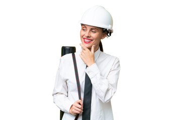 Young architect woman with helmet and holding blueprints over isolated background looking to the side and smiling