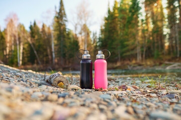metal thermo bottle on pebble beach of river  - obrazy, fototapety, plakaty