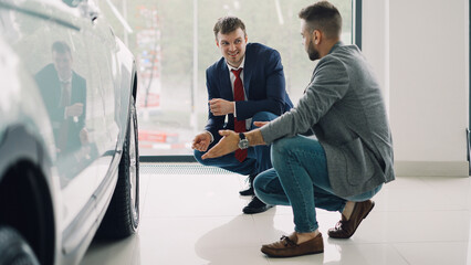 Two handsome men cheerful customer and friendly salesman are talking and gesturing discussing car model while squatting near expensive automobile. Buying and selling vehicles concept.