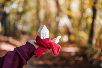 Miniature house in a red scarf in hand on an autumn background with yellow leaves. Thermal...