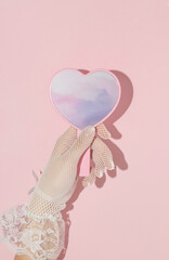 Valentines day creative layout with woman hand holding heart shaped mirror with pink clouds reflection on  pastel baby background. 80s, 90s retro romantic aesthetic love concept. Minimal fashion idea.