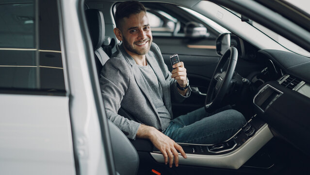 Close-up portrait of handsome bearded guy new car owner sitting inside beautiful automobile holding key fob and smiling looking at camera. Transportation and people concept.