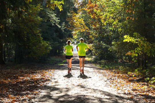 Two Runners Running Away From Camera On Sunny Fall Forest Unpaved Road.