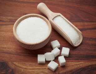 Miseczka i łopatka z białym cukrem, obok duże kostki białego cukru na drewnianym tle