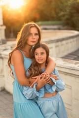 Portrait of mother and daughter in blue dresses with flowing long hair against the backdrop of sunset. The woman hugs and presses the girl to her. They are looking at the camera.