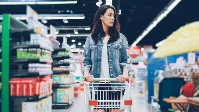 Young pertty brunette is walking in hypermarket and pushing shopping trolley going to buy food and looking around at shelves with products. People, consumerism and shops concept.