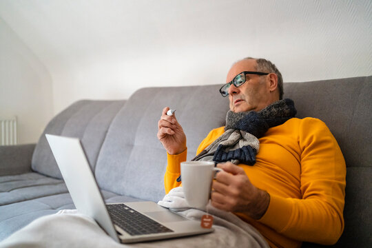 Man with laptop holding mug checking fever through thermometer on sofa