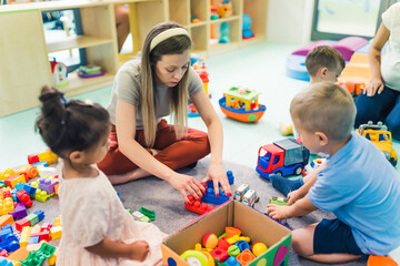 young blond teacher building with blocks for her beloved kids in the nursery, sitting on the floor numerous toys in the background. High quality photo