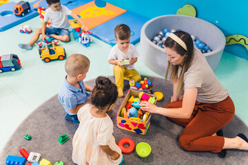 young Caucasian teacher sitting with her kids and playing with plastic toys, kindergarten and...