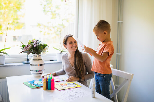 Smiling mother looking at son holding paper standing on chair