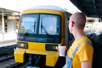 Transgender person with disposable coffee cup looking at train arriving on station