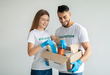 Happy activist volunteers packing food donations, putting cans and jars in cardboard box, standing over light wall