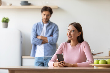 Offended sad millennial male looks at smiling female with smartphone at kitchen