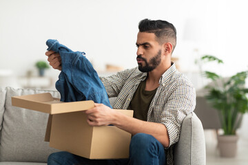 Upset young Arab guy unboxing carton package, receiving new clothes, unhappy about delivered item at home